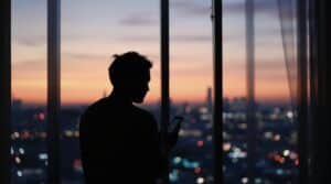 Silhouette d'un homme utilisant un téléphone devant une fenêtre avec vue sur la ville au crépuscule